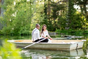 A happy couple, dressed in formal attire, smiles while rowing a small boat together on a calm lake surrounded by lush green trees.