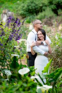 A couple in wedding attire embraces lovingly in a lush, flower-filled garden. The groom stands behind the bride, wrapping his arms around her as she closes her eyes and smiles peacefully.