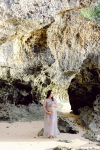 A pregnant woman in a flowing, sheer dress stands barefoot on sandy ground, surrounded by large rocky cliffs and natural cave formations, bathed in soft natural light.