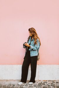 A woman with long hair stands against a pink wall, wearing a denim jacket, black pants, and white shoes. She is looking down at a camera in her hands and smiling slightly. Cobblestone pavement is visible at her feet.