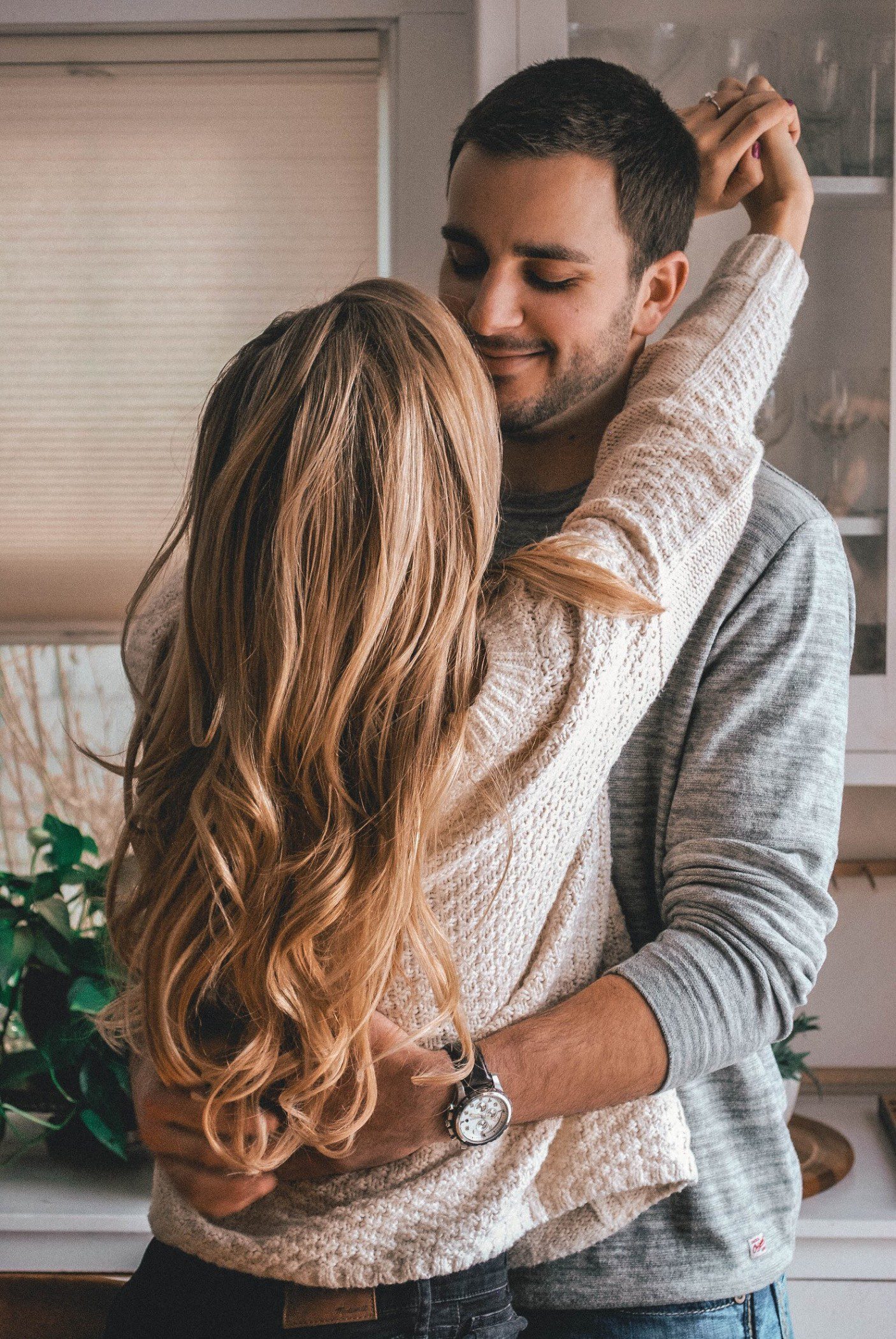 A couple embraces in a cozy kitchen, smiling warmly at each other. The woman has long, wavy blonde hair and wears a white sweater, while the man wears a gray sweater and holds her close.