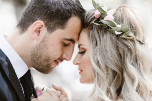 A bride and groom gently touch foreheads, eyes closed, sharing an intimate moment. The bride wears a floral crown and has wavy blonde hair; the groom is in a black suit and tie.