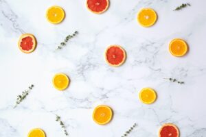 Slices of orange and grapefruit, along with sprigs of green herbs, are evenly spaced on a white marble surface, creating a bright, fresh, and minimalistic flat lay arrangement.
