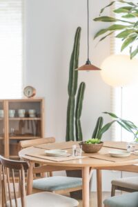 A bright, minimalist dining area with a wooden table set for three, a bowl of greens in the center, wooden chairs, tall green plants, a hanging pendant light, and a cabinet with dishes in the background.