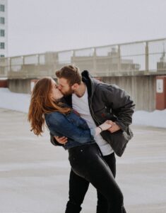 A young couple stands on a rooftop parking lot, leaning into a passionate kiss. The woman has long hair and wears a denim jacket, while the man has short hair and wears a gray jacket. Snow is visible on the ground behind them.
