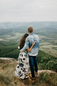 A couple stands on a grassy cliff, embracing as they look out over a vast, green valley and distant hills under a cloudy sky. The woman wears a floral dress; the man wears jeans and a denim jacket.