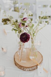Glass bottles with white and purple flowers are arranged on a wooden slice centerpiece, surrounded by small lit candles on a white tablecloth with white chairs in the background.