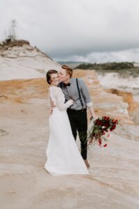 A groom kisses his smiling bride on the cheek while holding a bouquet of red and pink flowers. They stand on rocky terrain with cloudy skies and hills in the background.