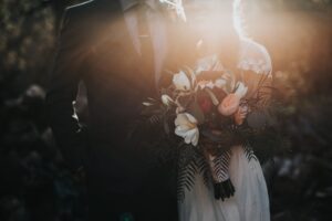 A bride in a white dress holding a bouquet of flowers stands beside a groom in a suit. Sunlight softly shines on them, creating a warm, romantic atmosphere. Their faces are partially obscured by the light.