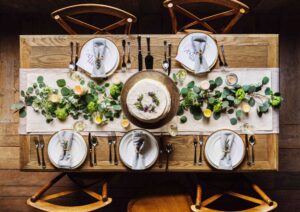 A wooden dining table set for four with white plates, gray napkins, silver cutlery, and name cards. A cake is in the center, surrounded by greenery, candles, and lemon slices as decoration.