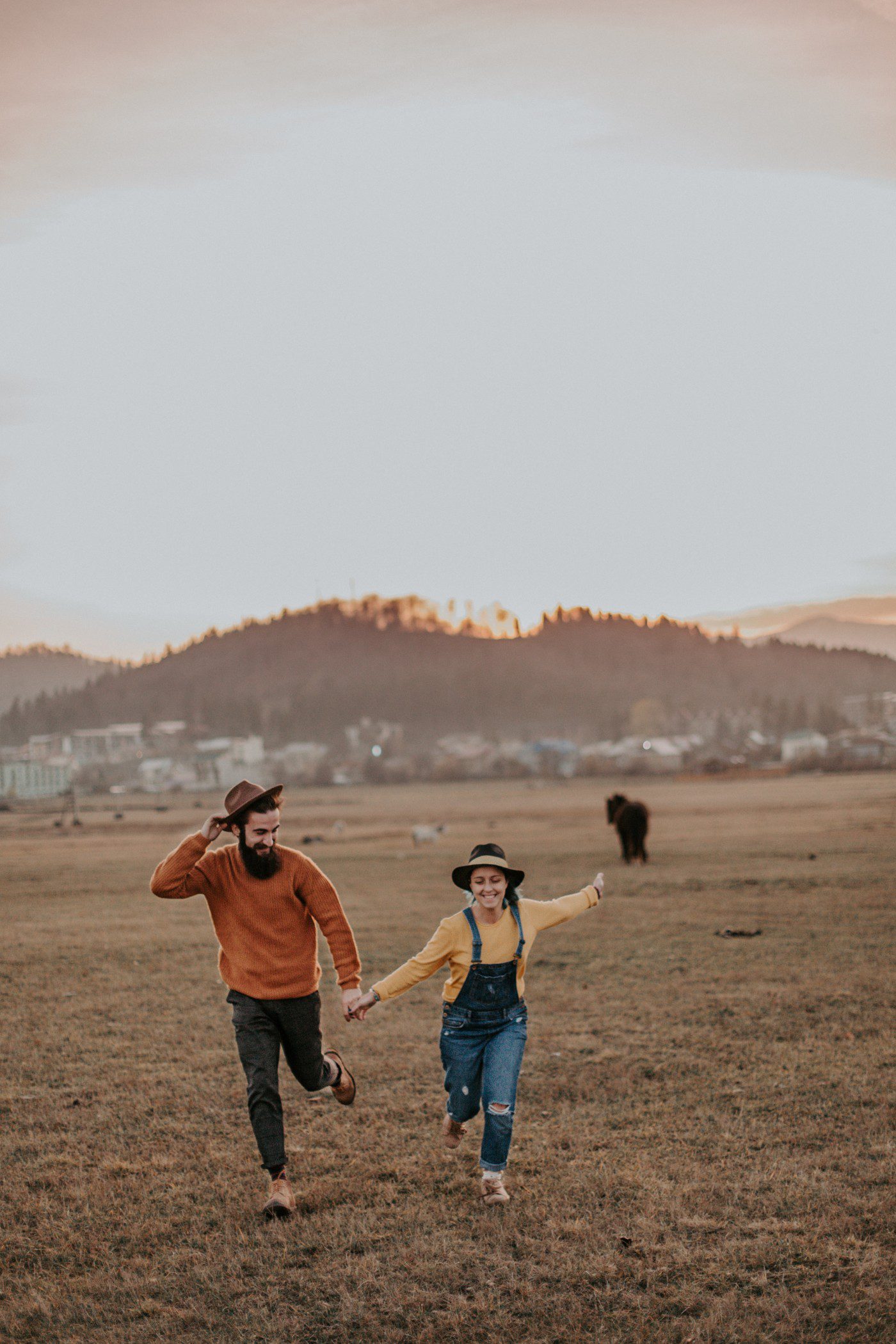 A smiling man and woman run hand-in-hand across a grassy field at sunset, both wearing hats and casual clothes. A distant hill, a few buildings, and a large animal appear in the background.