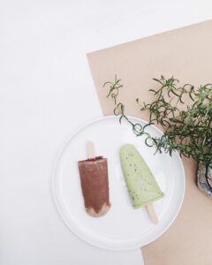 A white plate with two popsicles, one brown and one light green with black specks, is placed on a white surface next to a beige paper and green leafy plant.