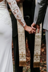 A bride and groom hold hands during their wedding ceremony, standing in front of an officiant wearing a decorative stole. The focus is on their joined hands and the details of the bride’s lace sleeve.