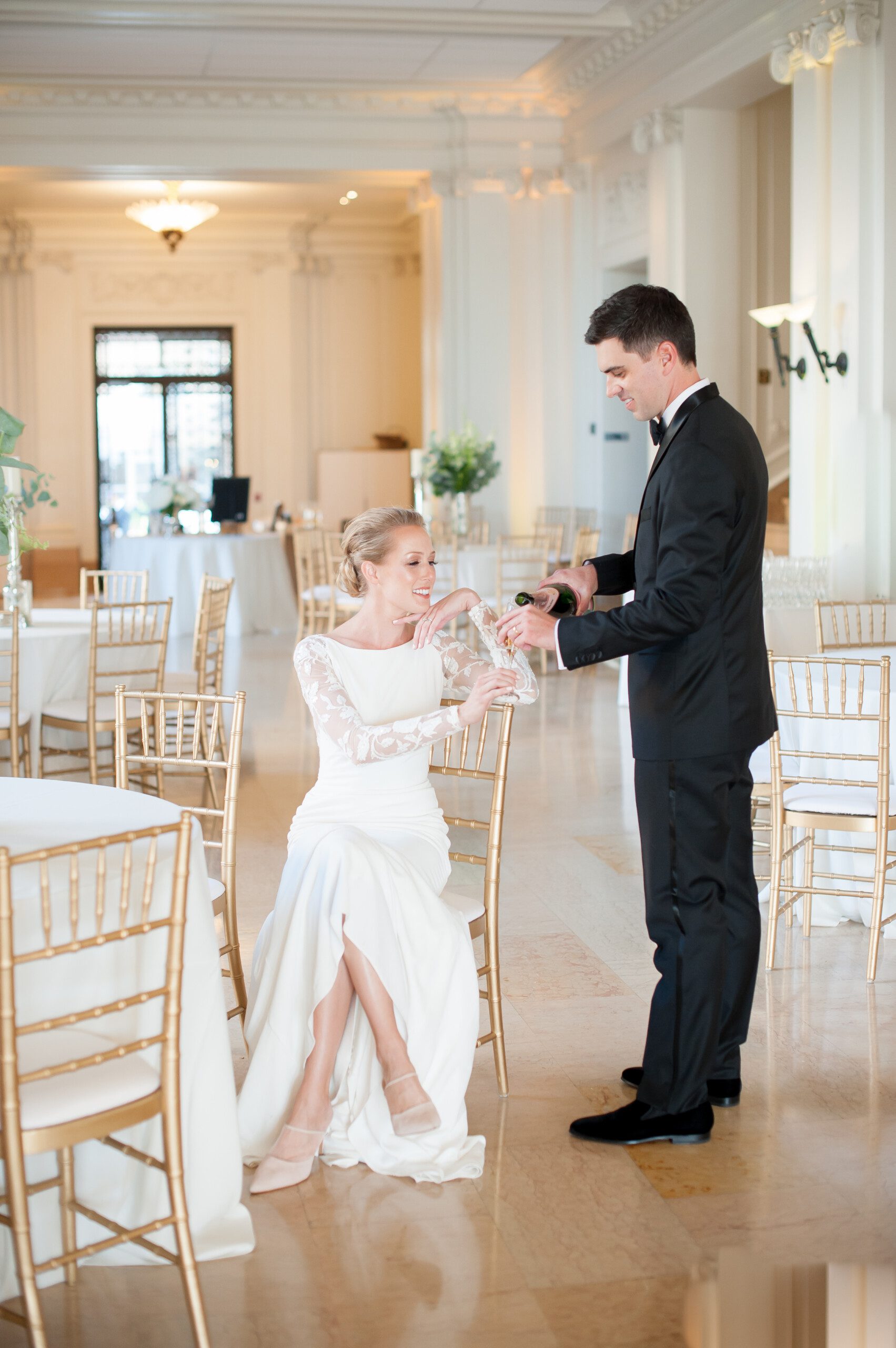 A bride in a white dress sits at a table while a groom in a black tuxedo pours her a drink in an elegant, bright reception hall with gold chairs and white décor.