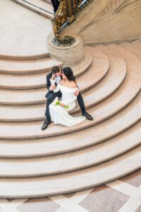 A bride in a white dress and a groom in a suit sit on marble steps, embracing and sharing a kiss. The bride holds a small bouquet of flowers. Ornate railings and stone architecture appear in the background.