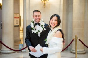A smiling bride and groom wearing leis stand together in formal attire inside an elegant building with columns and marble walls. The bride wears an off-the-shoulder white dress; the groom wears a black tuxedo.