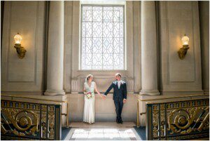A bride and groom stand hand in hand in front of a large ornate window between two columns in an elegant, sunlit hall with decorative railings and wall sconces.