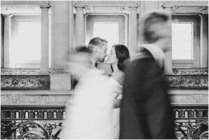 A couple kisses tenderly in focus on a grand indoor balcony, while two blurred figures pass by in the foreground, creating a sense of motion. The scene is in black and white.