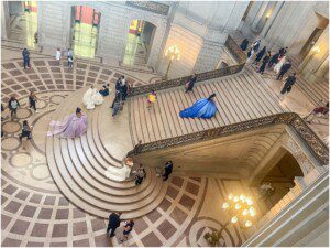 A grand staircase in an ornate building with several people posing for photos, some in elaborate gowns of white, purple, and blue. Other visitors walk or stand around the marble floor and steps. Bright chandeliers hang overhead.