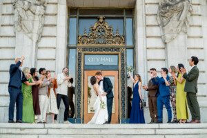 A couple kisses on the steps of City Hall, holding flowers, surrounded by friends and family cheering and clapping, dressed in formal and semi-formal attire.